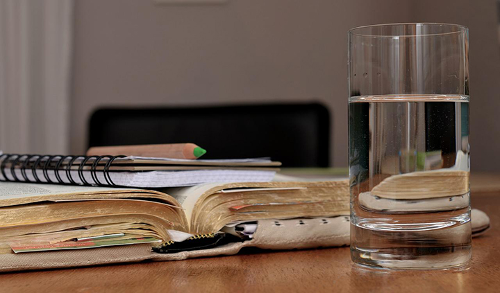 photo of a pencil resting on a spiral bound notebook resting on a large, yellowed cloth bound book, next to a glass of water